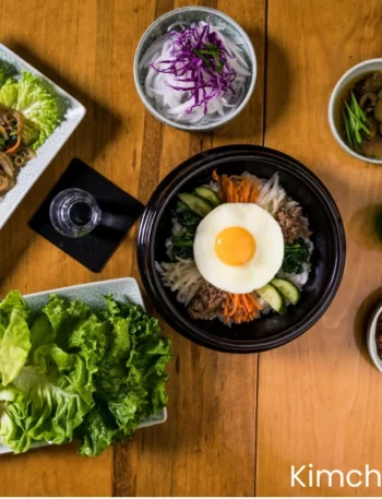 korean Bibimbap (Korean Mixed Rice with Meat and Assorted Vegetables) in a ceramic bowl , a water of glass, a Champaign bottle, some korean salids are placing on a wooden table