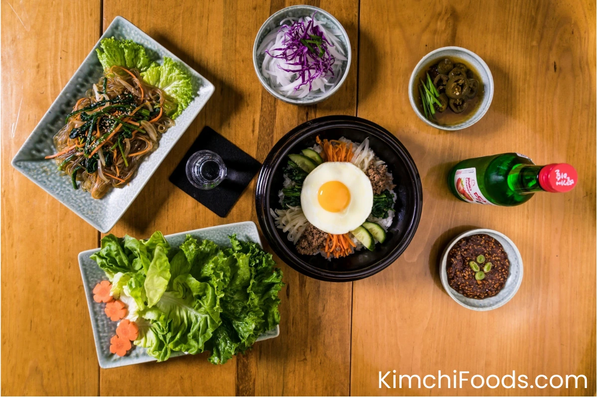 korean Bibimbap (Korean Mixed Rice with Meat and Assorted Vegetables) in a ceramic bowl , a water of glass, a Champaign bottle, some korean salids are placing on a wooden table